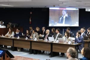 Foto colorida na posição paisagem mostra mesas onde um grupo de cerca de 15 pessoas estão sentadas. Eles participam da audiência de conciliação sobre as queimadas na Amazônia e no Pantanal. Ao fundo, em um telão, aparece a imagem do ministro Flávio Dino, que está falando ao microfone.