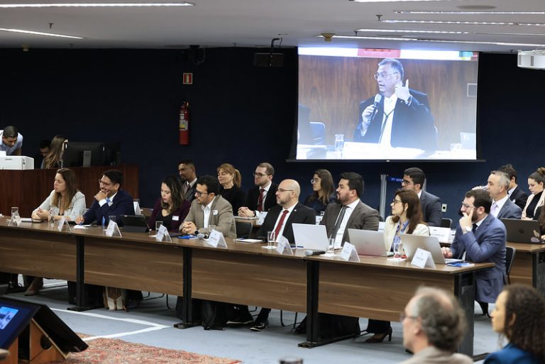 Foto colorida na posição paisagem mostra mesas onde um grupo de cerca de 15 pessoas estão sentadas. Eles participam da audiência de conciliação sobre as queimadas na Amazônia e no Pantanal. Ao fundo, em um telão, aparece a imagem do ministro Flávio Dino, que está falando ao microfone.