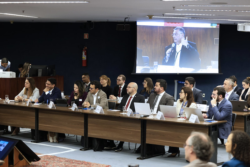Foto colorida na posição paisagem mostra mesas onde um grupo de cerca de 15 pessoas estão sentadas. Eles participam da audiência de conciliação sobre as queimadas na Amazônia e no Pantanal. Ao fundo, em um telão, aparece a imagem do ministro Flávio Dino, que está falando ao microfone.